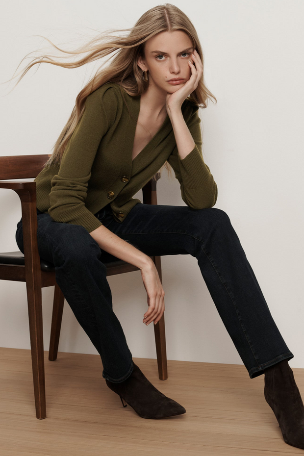 Woman sitting on a chair wearing an olive green cardigan and black pants.